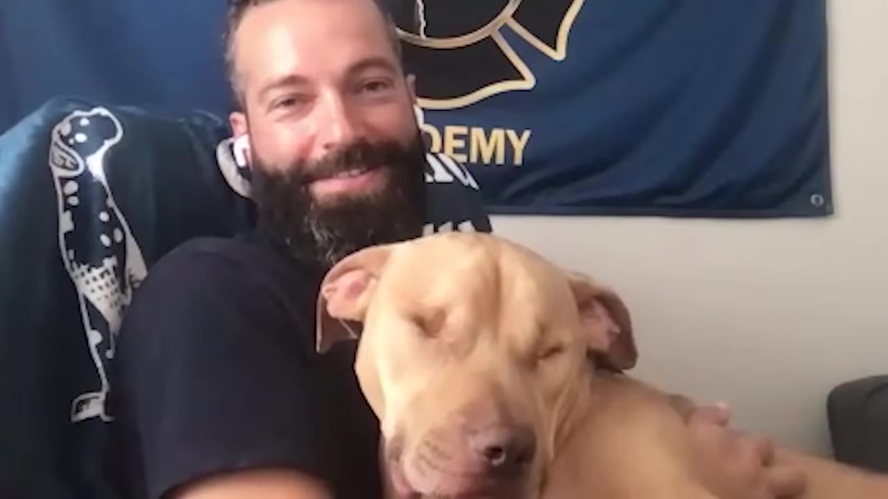 A smiling man with a beard holds a sleeping light brown dog close to his chest.