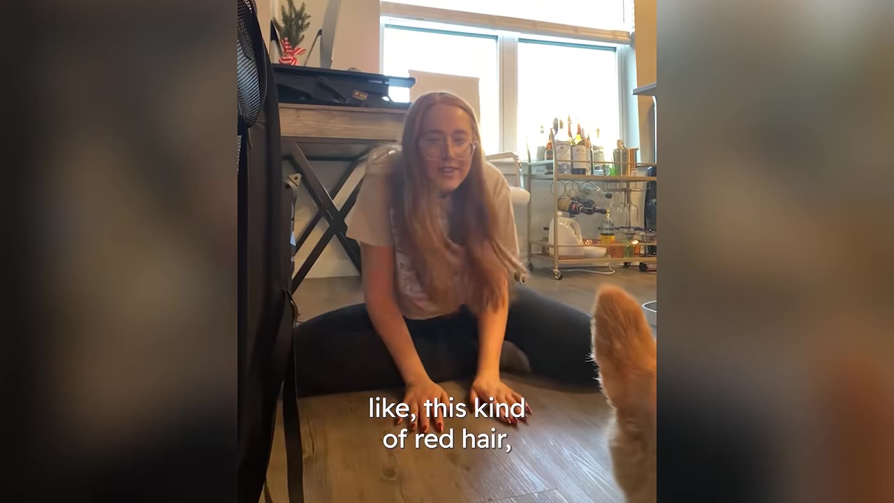 A woman with long red hair sits on the floor of her living room, looking towards a dog with matching reddish fur.