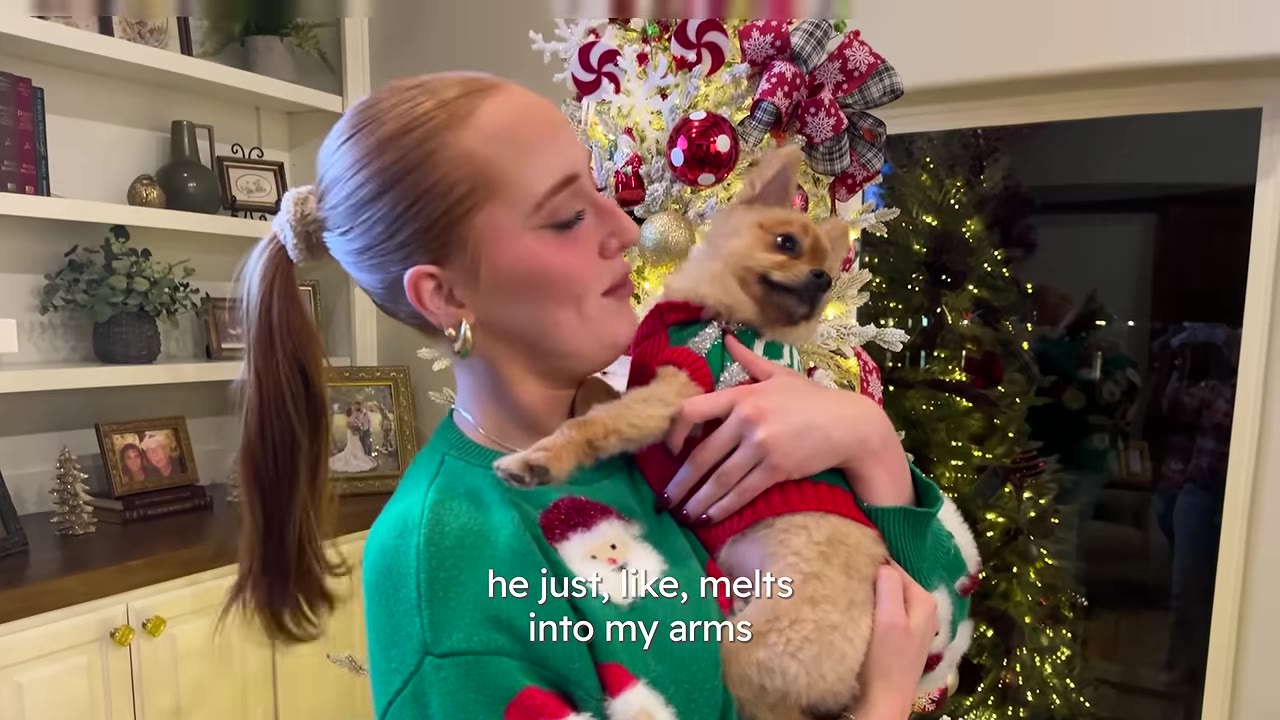 A woman with a blonde ponytail wearing a green Christmas sweater holds a small brown dog wearing a red and green sweater in her arms.