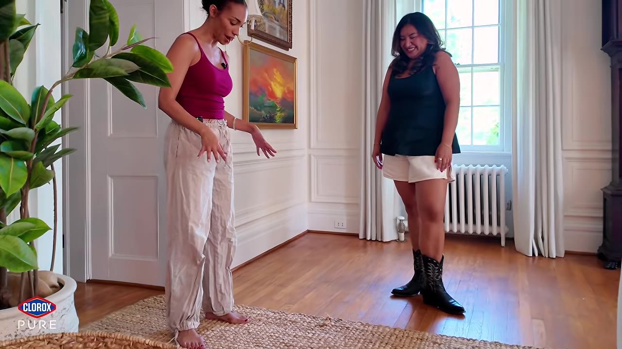 Two women stand in a room with hardwood floors and white walls, looking down at a long woven runner rug.