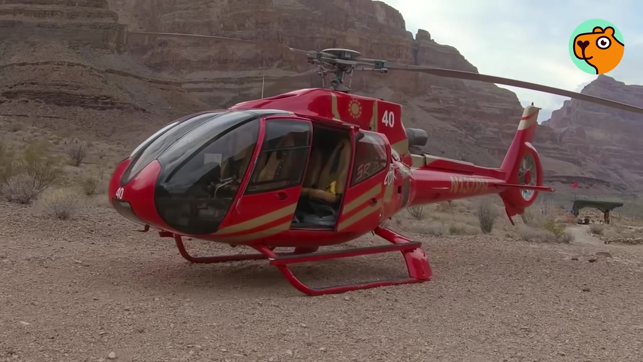 A red helicopter parked on dusty ground in a desert canyon in front of steep, rocky cliffs.