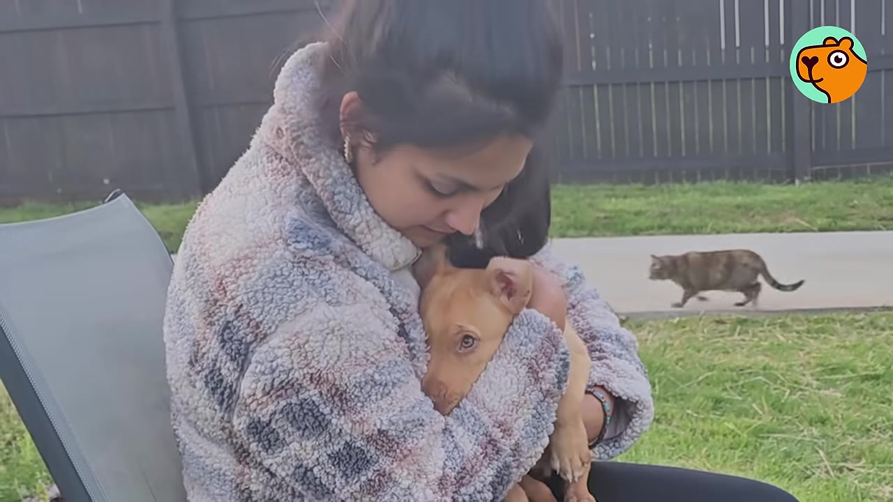 A woman wearing a cozy fleece jacket sitting outdoors and tenderly holding a small tan puppy in her arms.