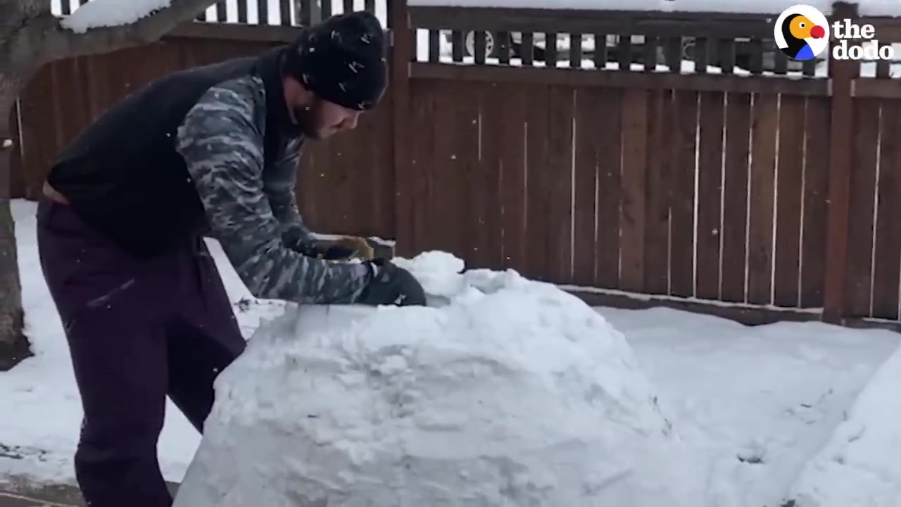A man wearing a beanie and winter clothes shaping a snow structure in a snowy backyard.