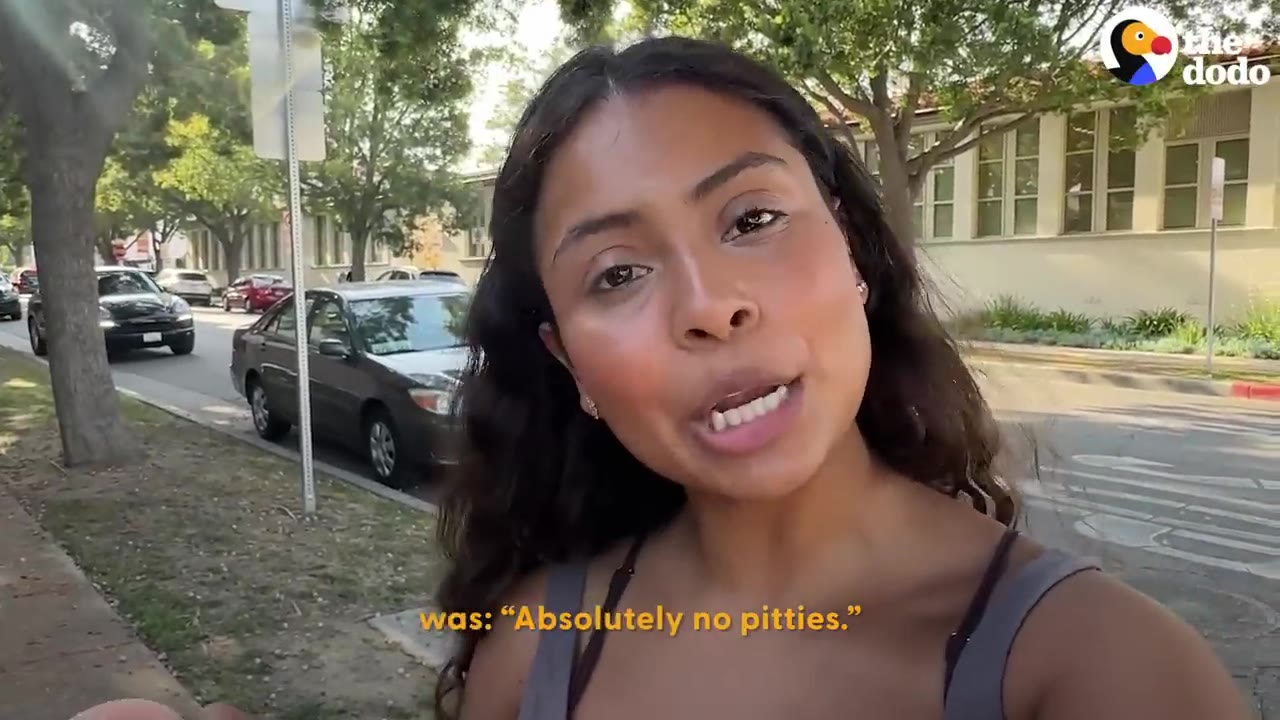 A young woman with long dark hair speaks to the camera on a sidewalk with cars parked in the background.