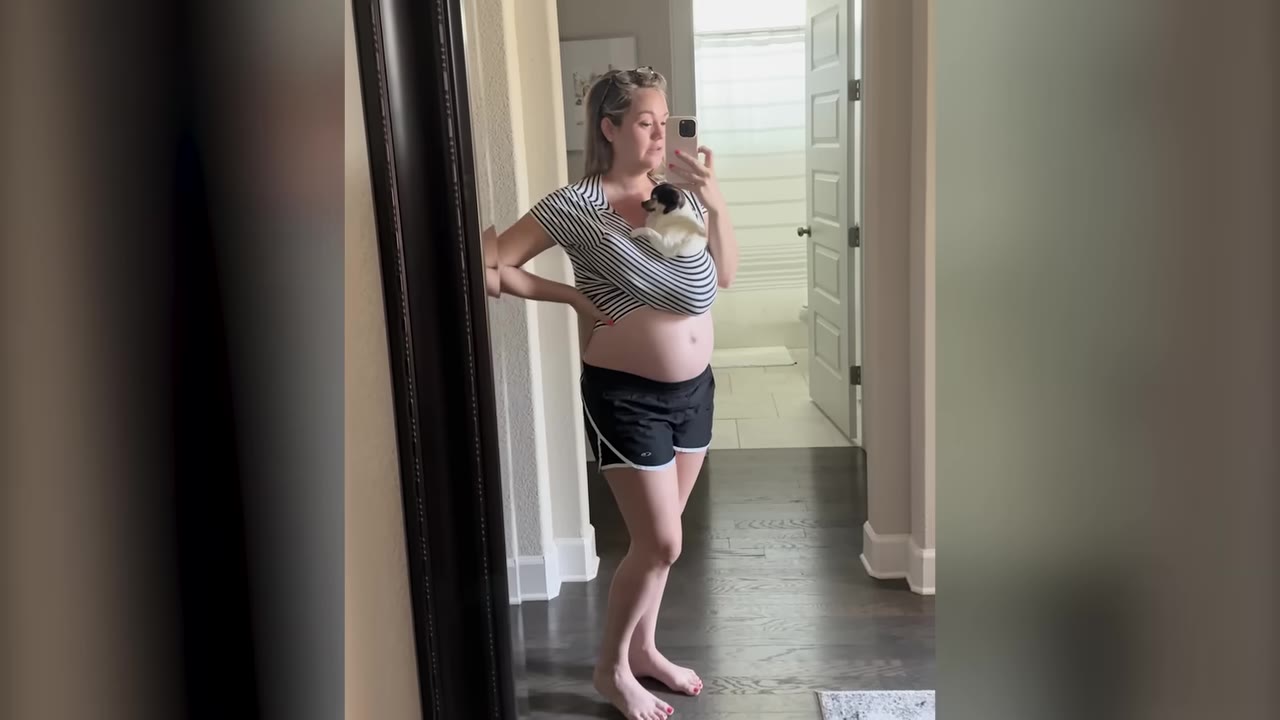 A pregnant woman taking a mirror selfie while carrying a small black and white dog tucked inside her shirt.