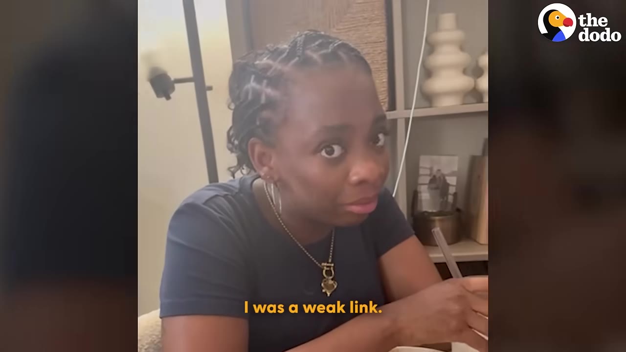 A woman with braided hair, wearing a dark blue t-shirt and a gold necklace, looks directly at the camera with a confessionary expression while sitting at a table.