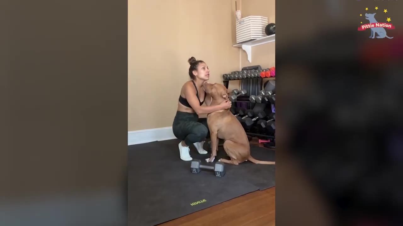 A woman crouching on an exercise mat in a home gym, holding and comforting a brown dog sitting beside her.