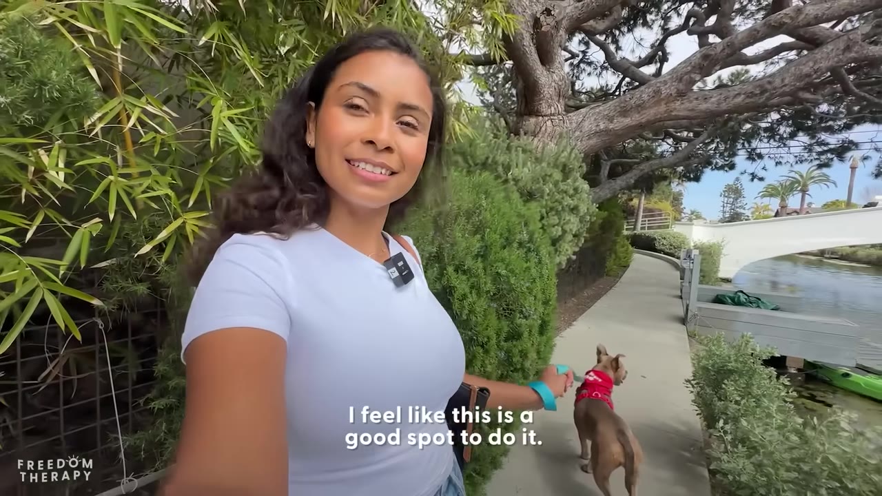 A woman in a white t-shirt smiles as she walks a small brown dog on a leash along a paved path next to a canal.