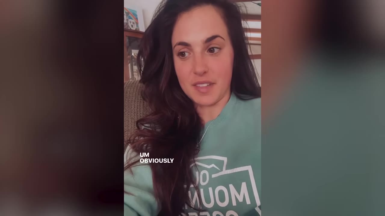 A woman with long dark hair wearing a light green sweatshirt speaks directly into the camera while recording a social media video.