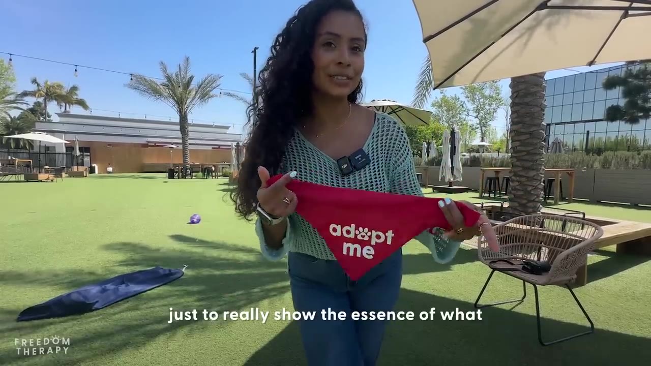 A woman with long curly hair stands on a green lawn holding up a red bandana that reads 'adopt me' with a paw print.
