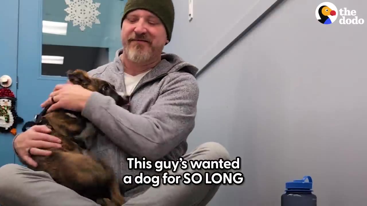 A man wearing a beanie and gray hoodie sitting cross-legged on the floor of an animal shelter room, lovingly hugging a small brindle puppy.