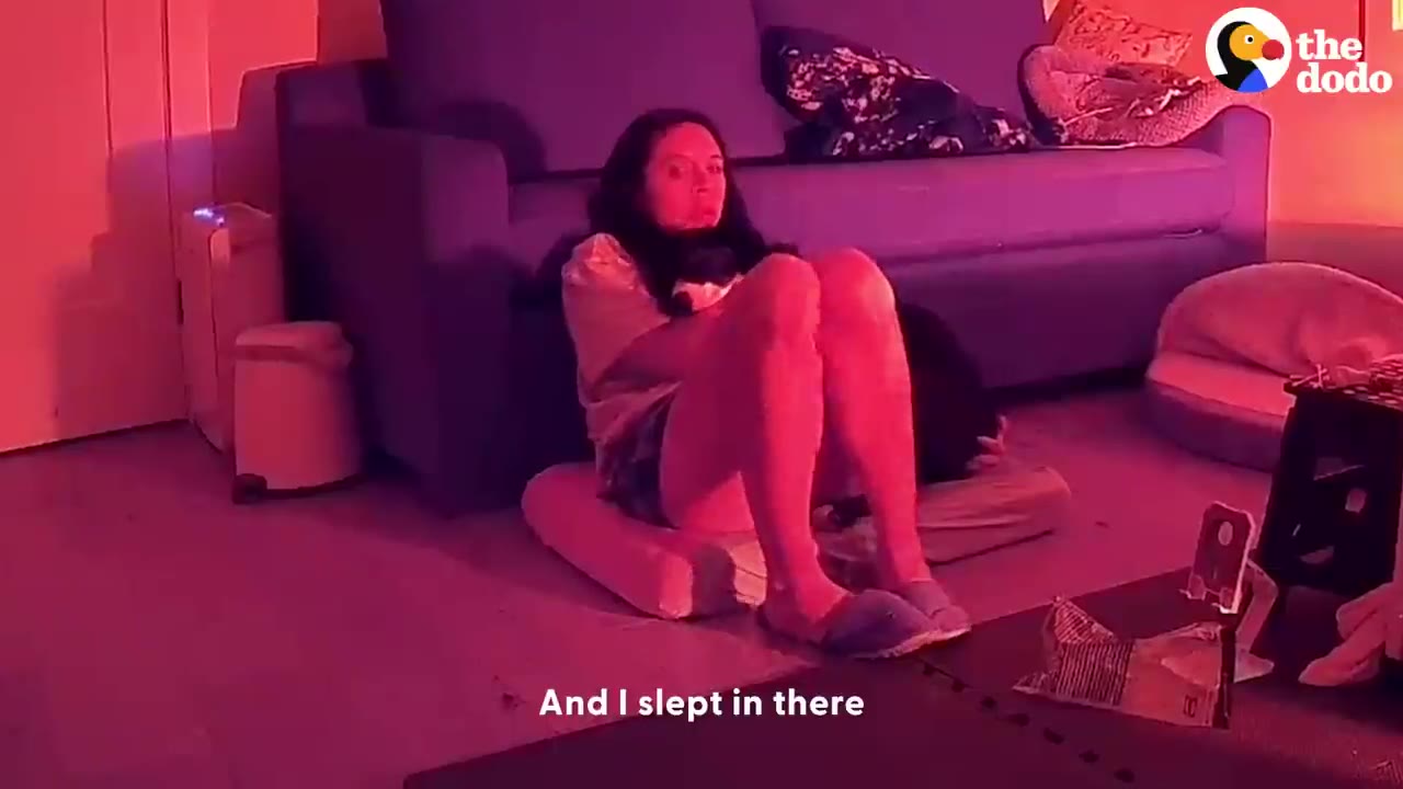 A woman sits on a cushion on the floor, leaning against a blue sofa in a dimly lit room with red lighting, looking tired.