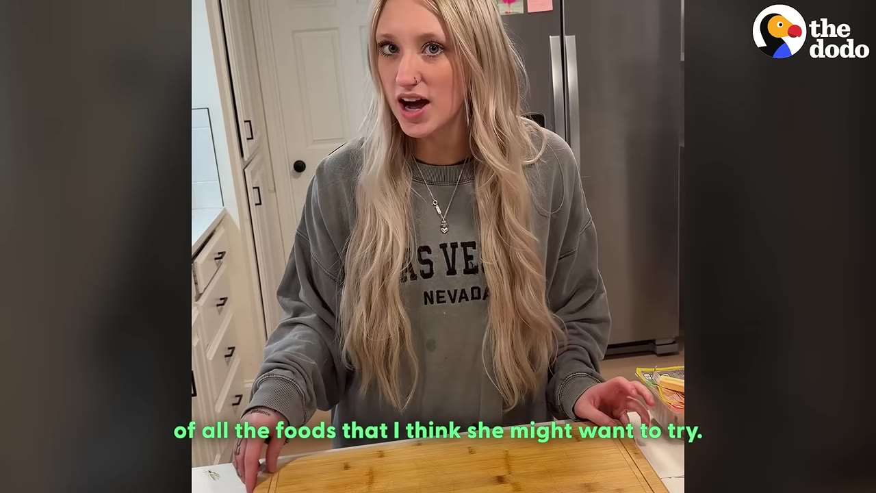 A young woman with long blonde hair stands in her kitchen behind a wooden cutting board, talking about preparing different foods for her dog.