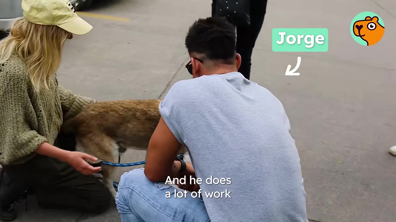 A man named Jorge crouching next to a woman who is petting a dog outdoors.