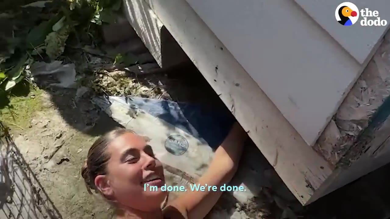A woman lying on the ground reaching under the edge of a house, looking up and smiling.