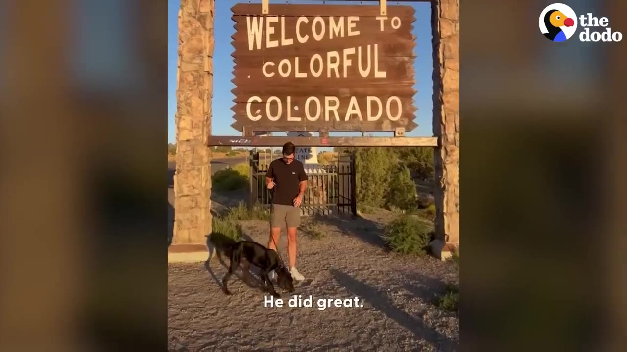 A man stands with a black dog on a leash in front of a large wooden sign reading Welcome to Colorful Colorado.