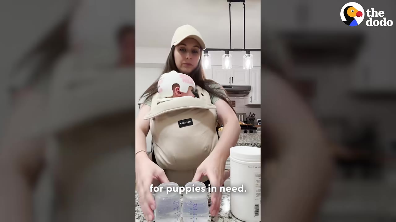 A woman wearing a baseball cap and a baby carrier with an infant inside prepares two small bottles on a kitchen counter.