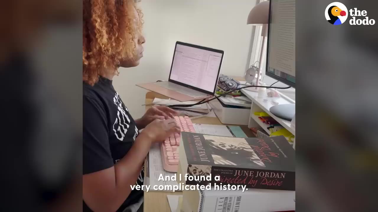 A woman with curly hair sitting at a desk typing on a pink keyboard surrounded by books and laptop screens.