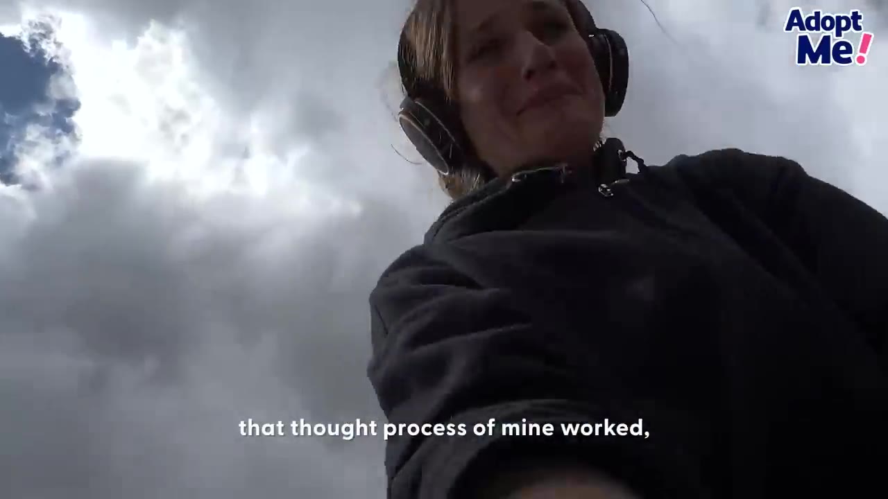 A woman wearing headphones and a dark hoodie smiles from a low angle against a cloudy sky.
