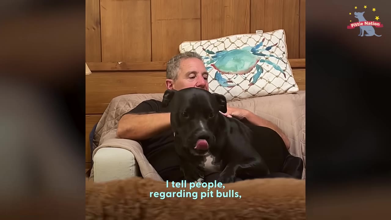 A man sits in an armchair, holding and petting a black dog resting on his lap.