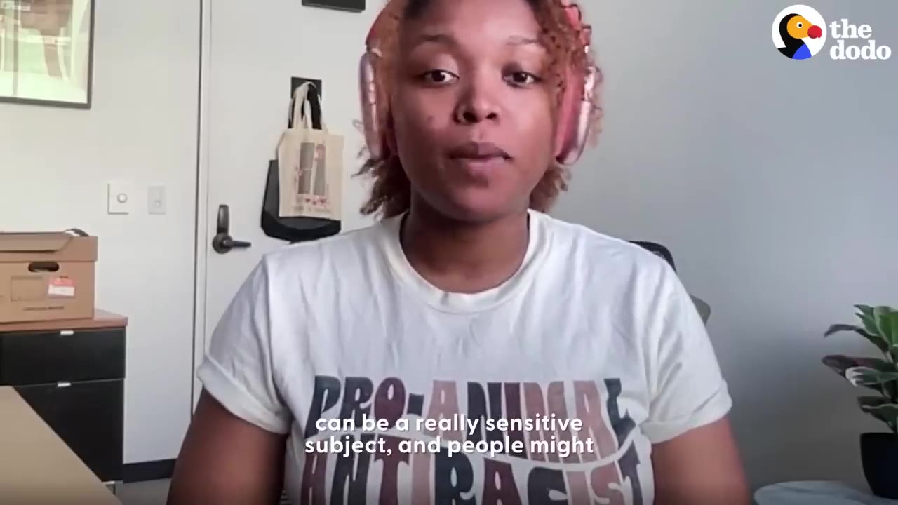A woman wearing headphones and a white t-shirt speaking directly to the camera indoors.