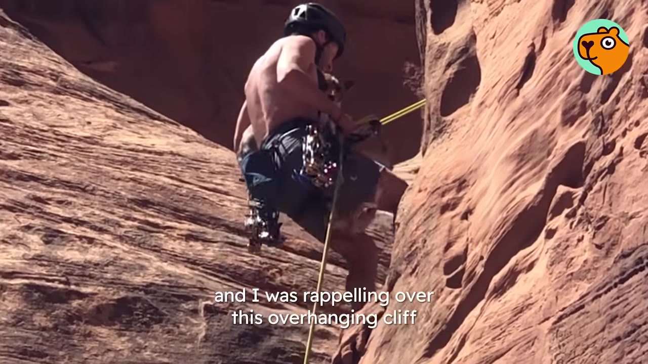 A shirtless man wearing a climbing helmet lowers himself down a steep red rock cliff edge on a yellow rappel rope.