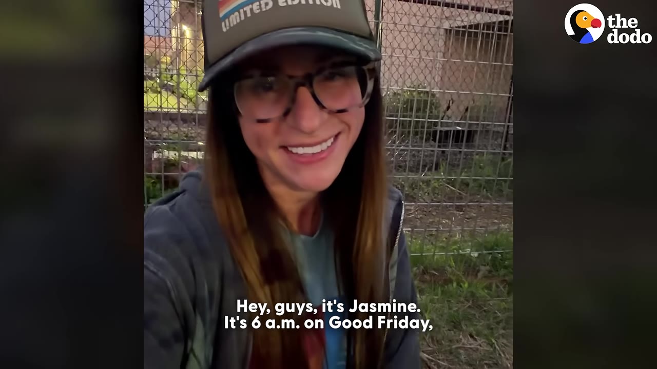 A close-up selfie of Jasmine wearing a baseball cap and glasses, smiling in front of a wire fence in the early morning light.