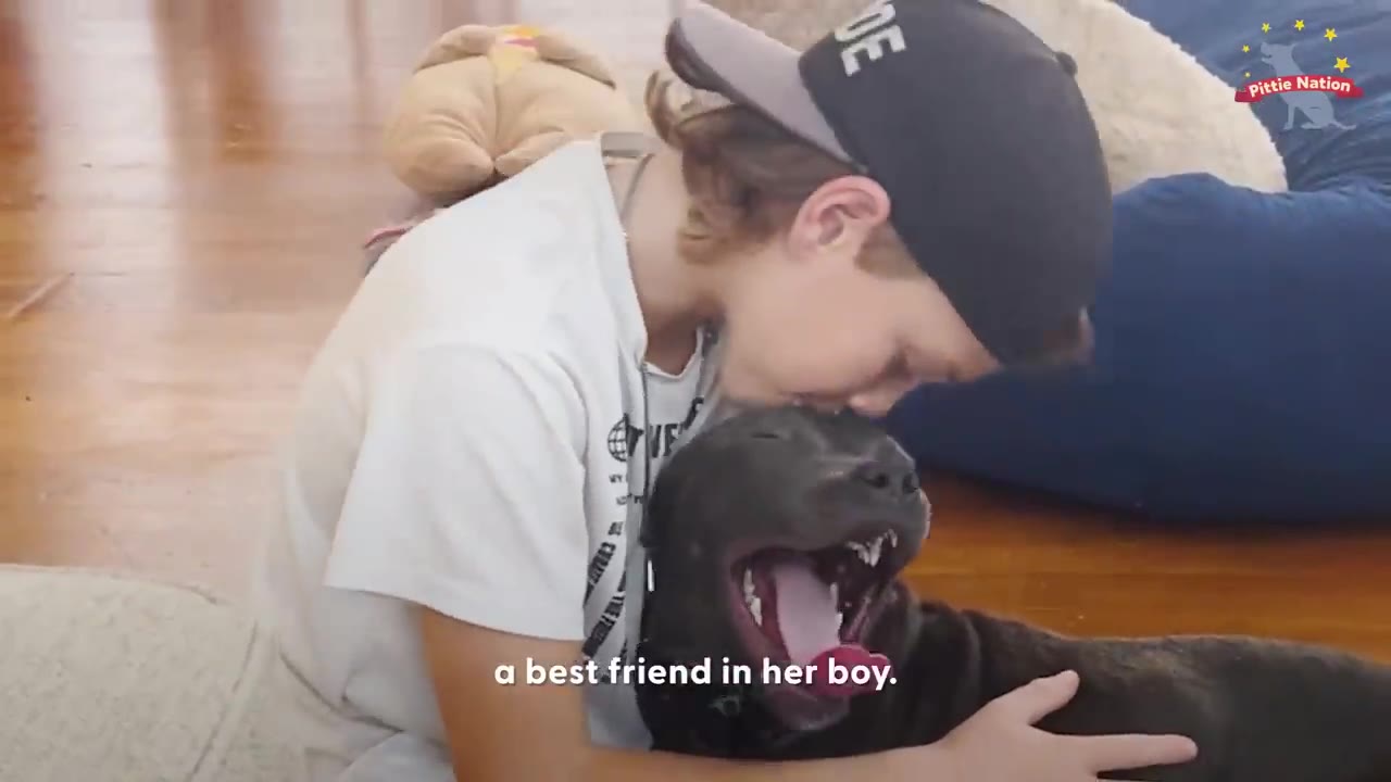 A boy wearing a backwards baseball cap hugs and kisses a black puppy on the head indoors.