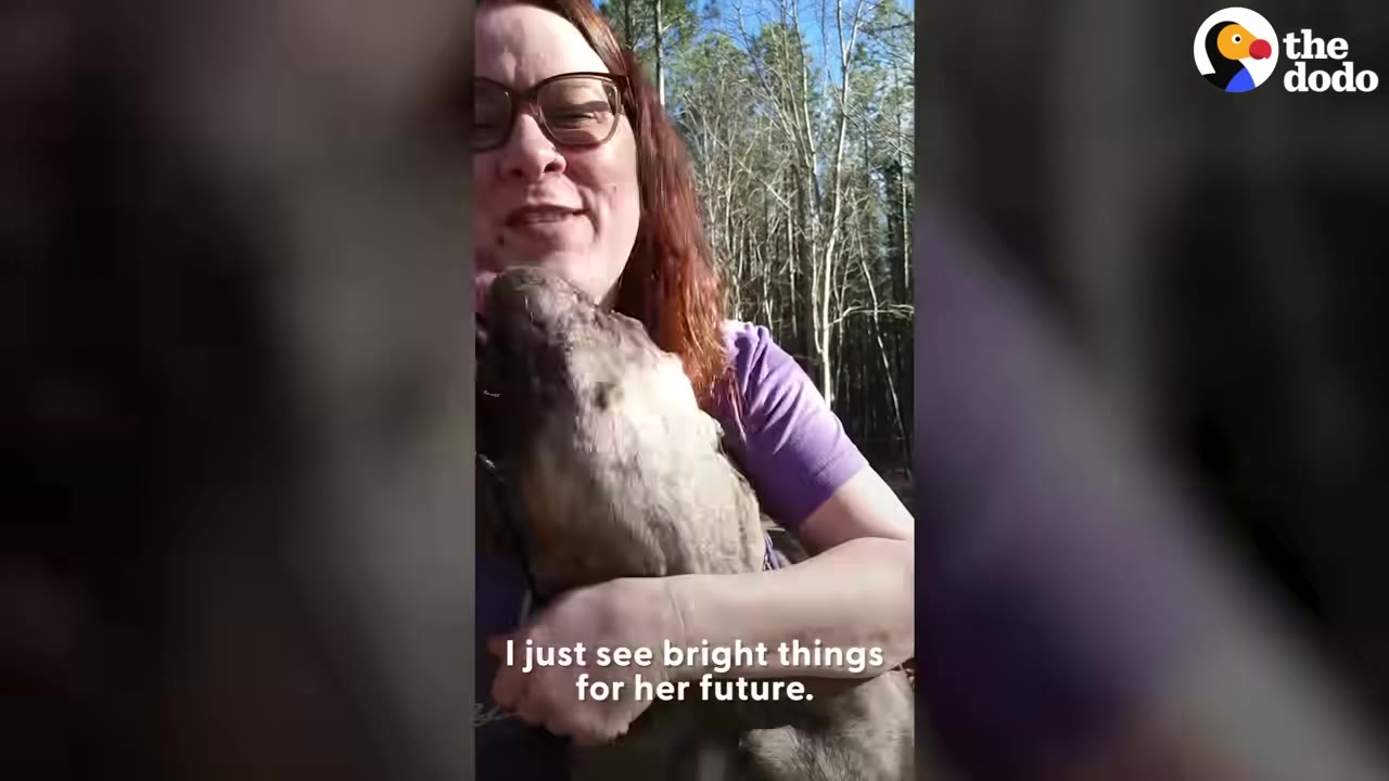 A woman wearing glasses and a purple shirt smiling while holding and cuddling a brindle dog outdoors.