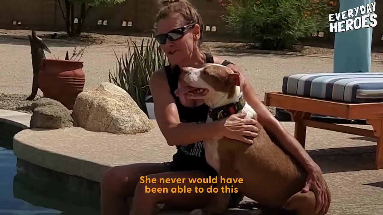 A woman wearing sunglasses sitting poolside and petting a brown and white dog sitting beside her.