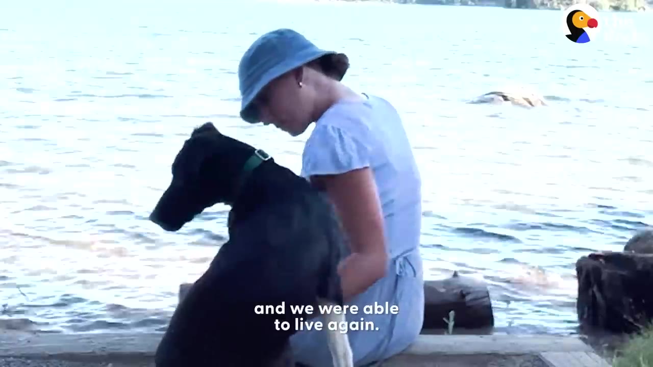 A woman in a blue bucket hat sitting by the water next to a black dog.