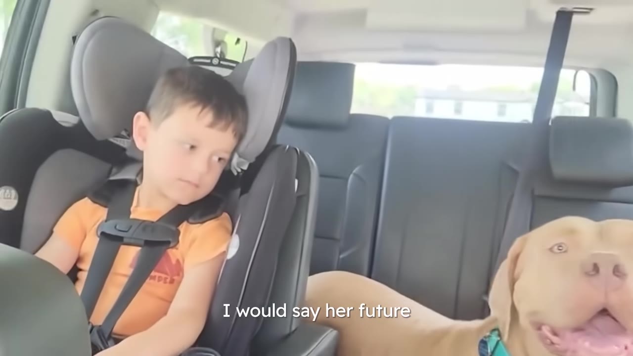 A happy brown dog sitting in the back seat of a car next to a young boy in a car seat.