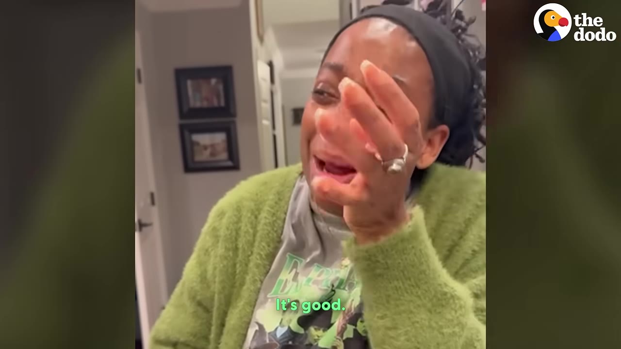 A close-up of a woman wearing a green cardigan and a black headband, crying with happy tears and covering her face with her hand.