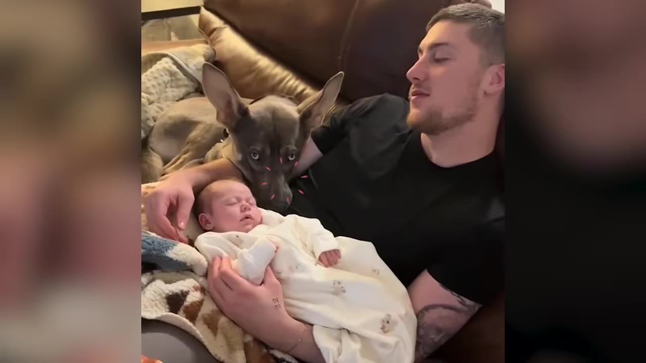 A dog rests its head on a couch behind a man holding a sleeping baby in a white onesie.