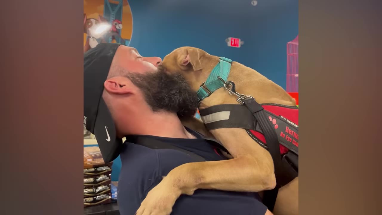 A man with a beard and a black cap holds a tan dog wearing a red harness close to his face in an affectionate embrace.