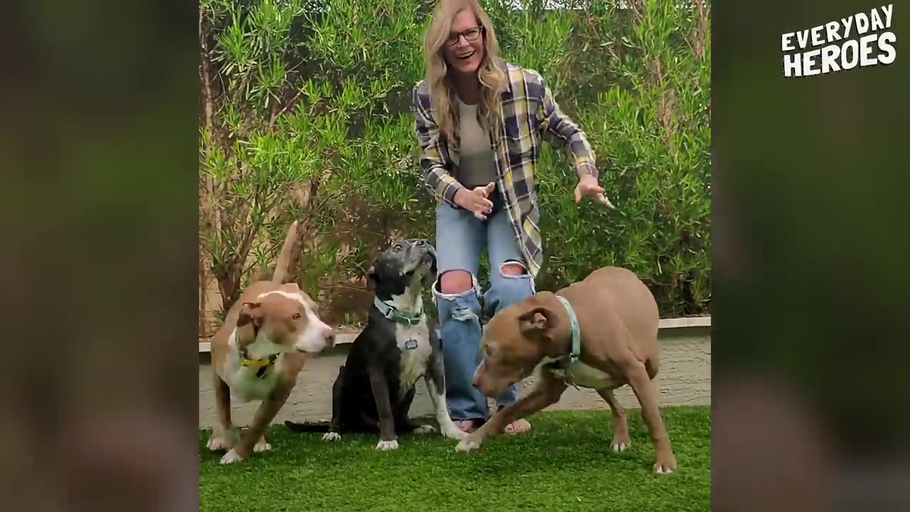 A smiling woman standing outdoors in a yard surrounded by three dogs.